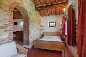 Una habitación con cama, una silla de mimbre, pared de piedra, techo de madera y una ventana.
