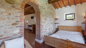 Une pièce avec un mur en pierre, un plafond en bois, un lit, une fenêtre et une porte donnant sur une autre pièce.