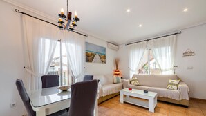Una sala de estar con una mesa de comedor, sillas, un sofá y una mesa de centro. Hay una araña, un cuadro en la pared y un equipo de aire acondicionado.