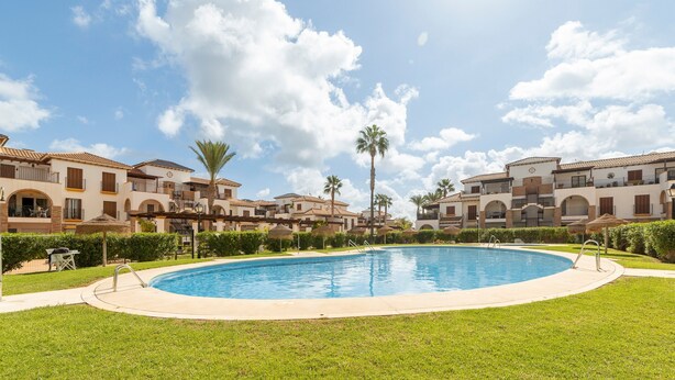 Una piscina rodeada de un césped cuidado, con edificios de varios pisos que tienen balcones y palmeras al fondo.