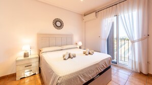 Un dormitorio con cama, mesitas de noche, un espejo montado en la pared y vistas a través de una ventana con cortinas.