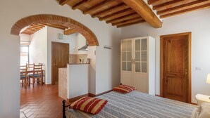 A bedroom with a bed, wooden ceiling beams, a brick archway, a wardrobe, and a kitchen area. - Podere Papèra - 'Volpe Flat' Apartment with Mountain View, Wi-Fi and Air Conditioning (San Casciano in Val di Pesa)