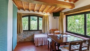 A room with a stone wall, wooden ceiling beams, a bed with a pink bedspread, a table with chairs, and large windows with curtains. - Podere Papèra - 'Cavallo Flat' Apartment with Mountain View, Wi-Fi and Air Conditioning (San Casciano in Val di Pesa)