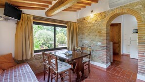 A room with a stone wall, wooden ceiling beams, a dining table with chairs, a television, and a window with curtains. - Podere Papèra - 'Cavallo Flat' Apartment with Mountain View, Wi-Fi and Air Conditioning (San Casciano in Val di Pesa)