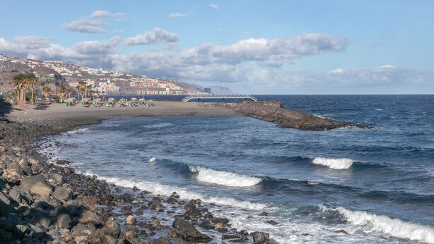 Una playa costera con costa rocosa, un muelle y edificios al fondo.