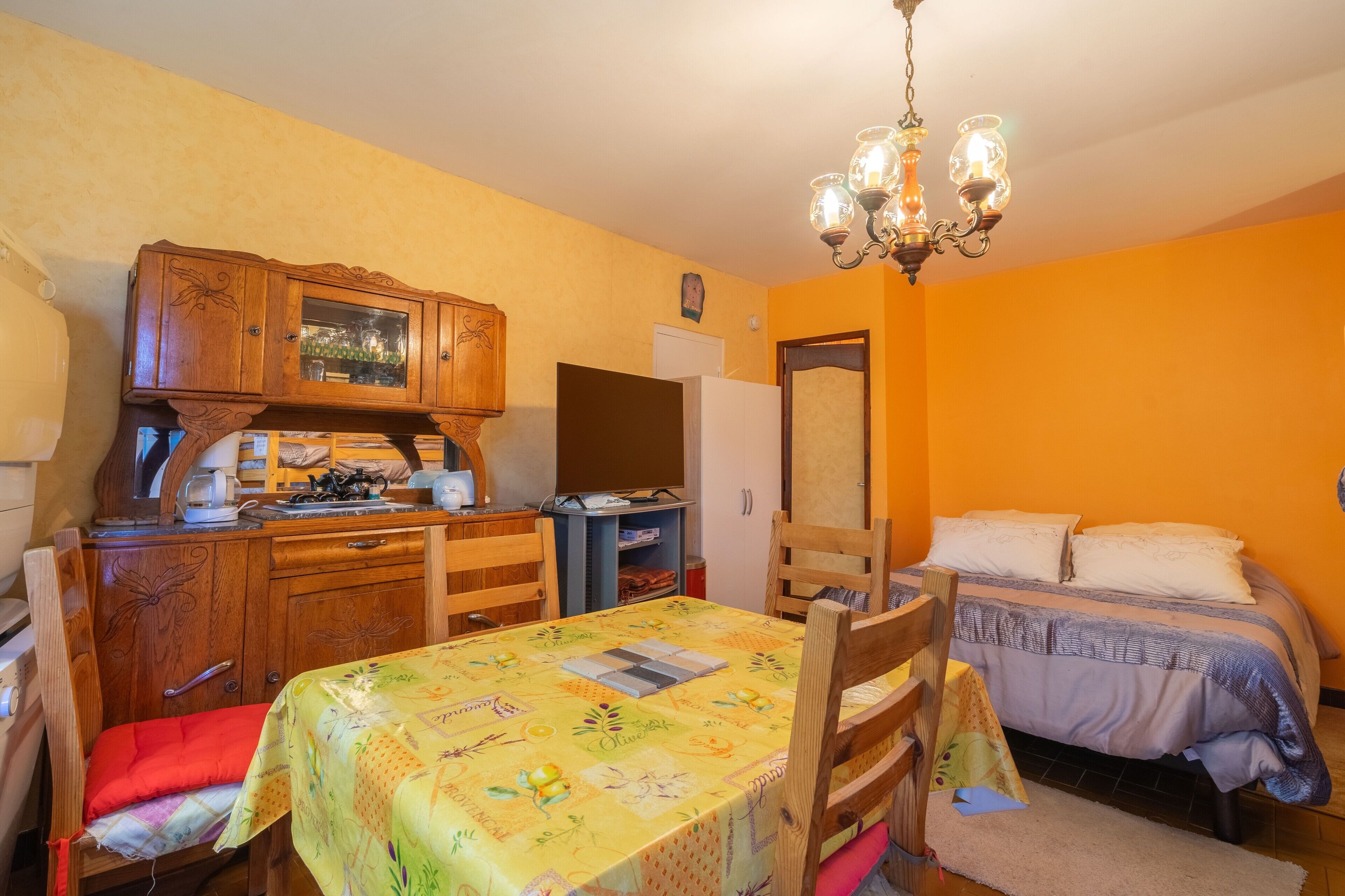 A compact room with a dining table, chairs, a bed, a wooden cabinet, and a chandelier.