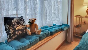 Ein Zimmer mit Fensterbank, einem Teddybären, einem schwarzen Kissen mit Baummotiv und einer Lampe auf einem Beistelltisch.