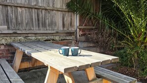 Ein Holztisch mit Picknick-Bestuhung, belegt mit zwei blauen Tassen, vor einer Ziegelwand und umgeben von Grünpflanzen.
