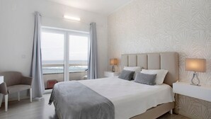 A hotel room with a large bed, a chair, a desk, a nightstand, and a view of the ocean.