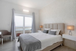 A hotel room with a large bed, a chair, a desk, a nightstand, and a view of the ocean.