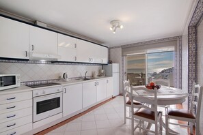 A modern kitchen with white cabinets, a microwave, stove, and oven. There is a dining table with chairs and a view of the sea through a glass door. - Apartment 'Apartamento Varandas Do Mar' with Sea View, Wi-Fi and Air Conditioning (Albufeira)