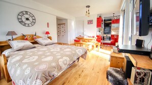 Quarto com cama, mesas de cabeceira, piso de madeira, área de cozinha com armários vermelhos e uma televisão.