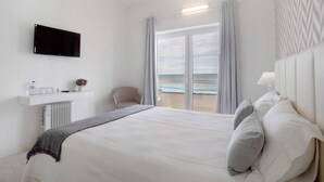 Habitación de hotel con cama, televisor, una silla, ventana con cortinas y vista al mar.