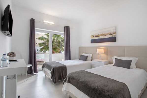 Une chambre d’hôtel avec deux lits, un bureau, une télévision, un tableau et une vue sur des palmiers.