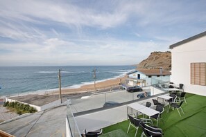 Un balcon avec vue sur la plage et l’océan, des sièges d’extérieur et un bâtiment doté d’un toit en pente.