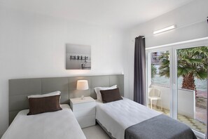 Quarto de hotel com duas camas, mesa de cabeceira com abajur, vista para o mar e varanda com cadeira.
