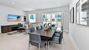 A modern dining area with a long table, multiple chairs, a dark wooden table, and a large window with curtains. - Modern, Luxury 1st Floor Condo Near Disney (Reunion)