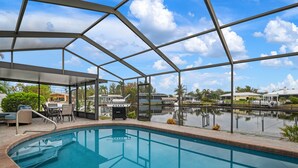 A pool area with a covered seating area, a grill, and a view of a lake and buildings in the background. - ⚓Sandys Coral Retreat! Canal Front, Pool, Hot Tub & More⚓ (Bradenton)