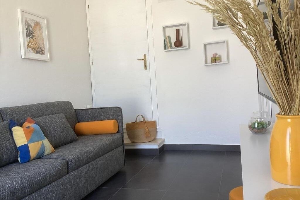 Ein modernes Wohnzimmer mit einem grauen Sofa, einem farbenfrohen Kissen, einem gewebten Korb und einer gelben Vase mit Trockenpflanzen.