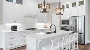 Una cucina moderna con mobili bianchi, un'isola centrale con sgabelli da bar, elettrodomestici in acciaio inossidabile e lampade a sospensione dalle forme geometriche.