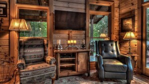 L'intérieur chaleureux d'un chalet avec un fauteuil en cuir, un canapé à motifs, un bureau en bois avec une télévision et une grande fenêtre donnant sur un paysage verdoyant.