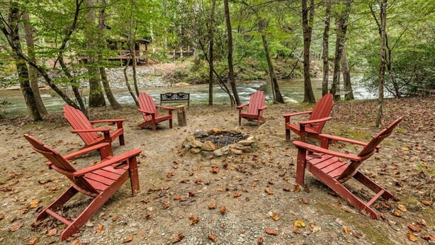 Un terrain de camping avec quatre chaises Adirondack rouges disposées autour d’un foyer central, le tout étant entouré d’une zone boisée, avec une rivière en arrière-plan.