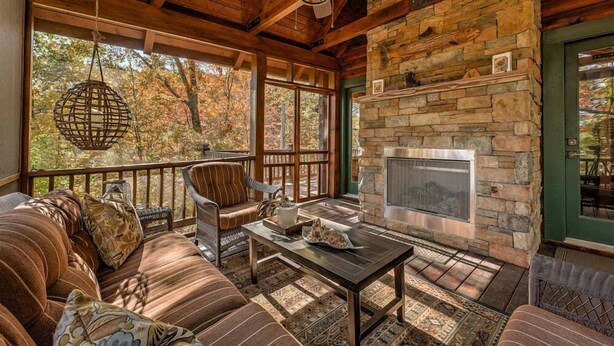 L’intérieur chaleureux d’un chalet avec une cheminée en pierre, des meubles en bois et de grandes fenêtres donnant sur une forêt.