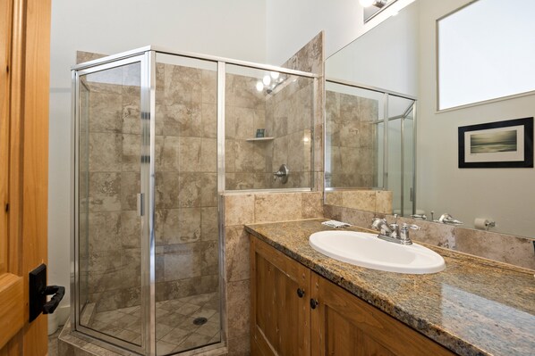 A modern bathroom with a glass-enclosed shower, a granite countertop sink, wooden cabinets, and a large mirror.