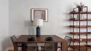 Een eethoek met een houten tafel, vier stoelen met gaasframe, een zwarte lamp en een boekenkast met een plant.