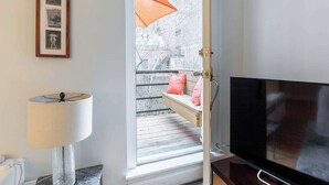 Een kamer met een televisie, een raam met uitzicht op een balkon, een lijst met een foto aan de muur en een lamp.