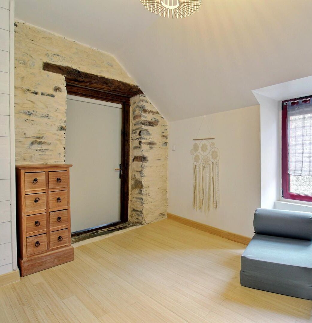 A room with a stone wall, a wooden dresser, a gray sofa, and a dreamcatcher on the wall.