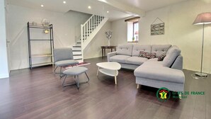 A modern living room with a grey sofa, a small round table, and a chair. There's a staircase in the background and a lamp on the right. - Maison de la Vilaine (SAINT DOLAY)