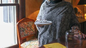 Una persona de pie junto a una ventana, con una silla y una mesa que sostiene una bandeja y un recipiente de vidrio.