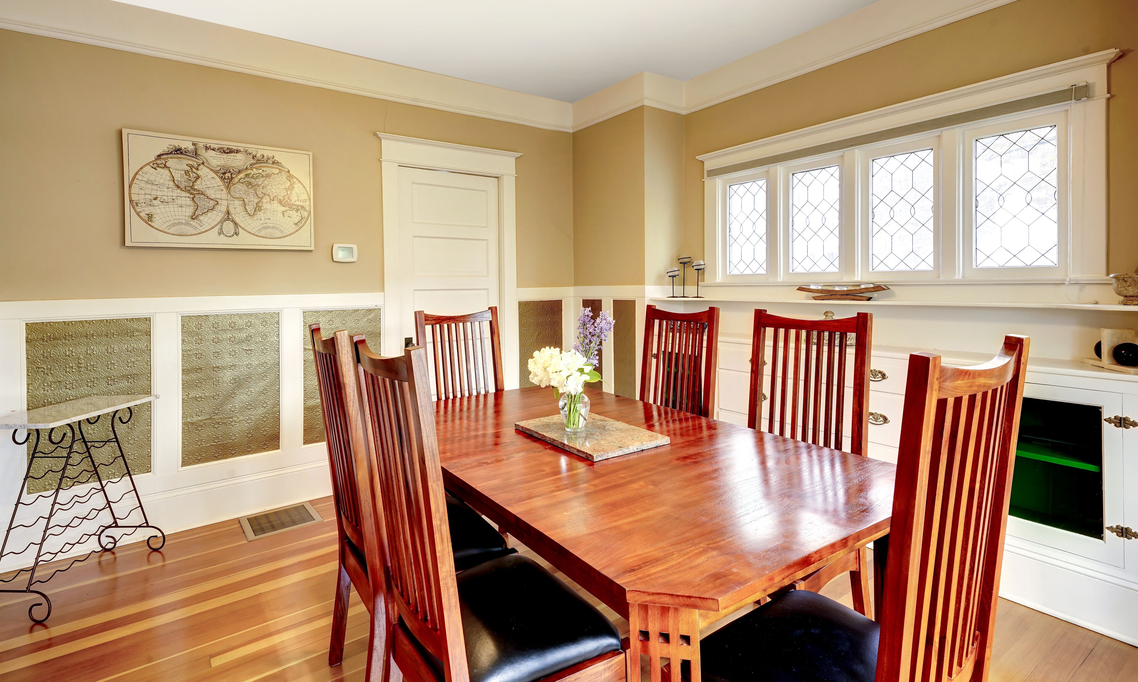 A dining room with a wooden table, six chairs, a vase with flowers, and a world map on the wall.
