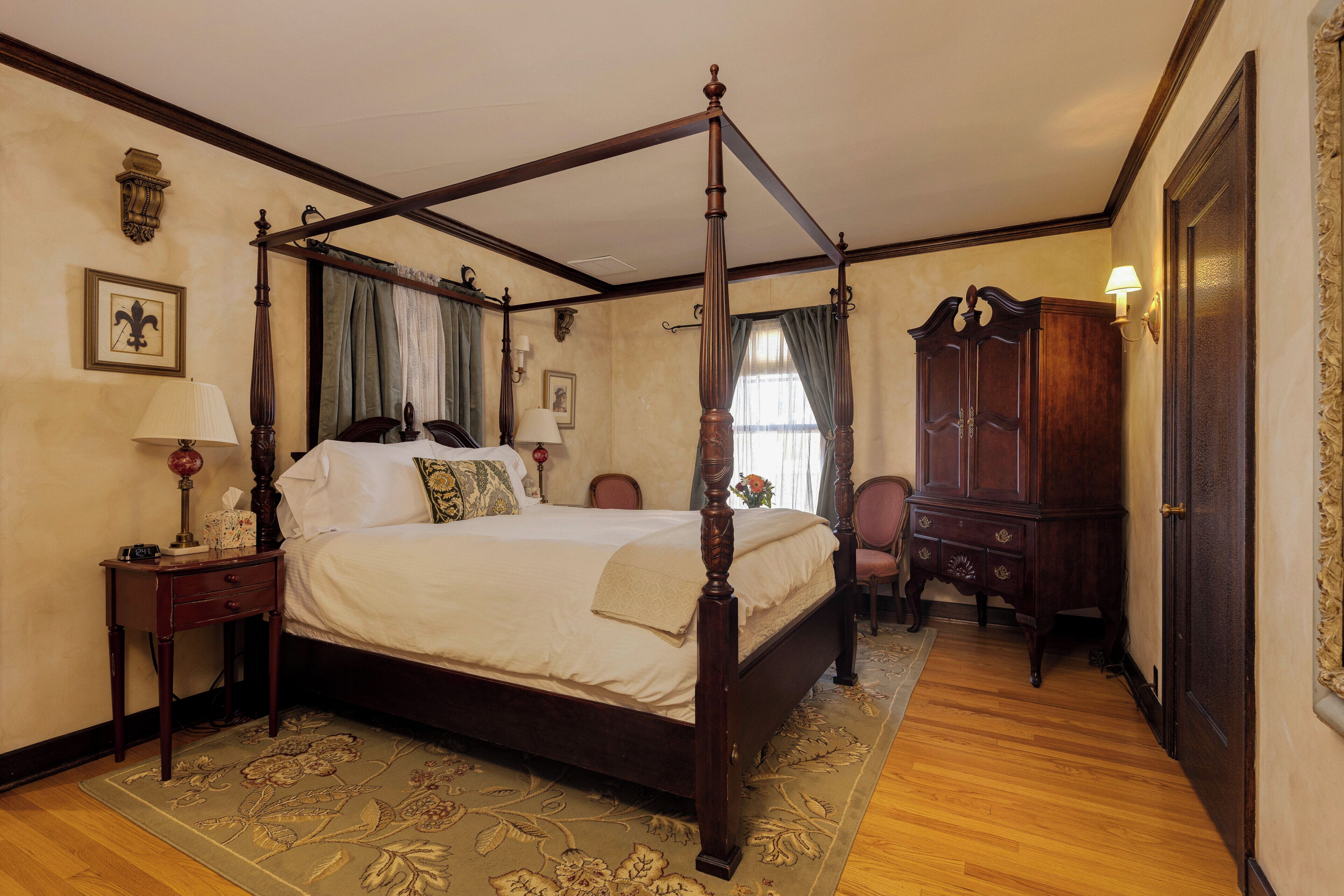 A four-poster bed with a wooden frame, a nightstand with a lamp, a chair, and a wooden cabinet in a room with wooden flooring and a floral-patterned rug.