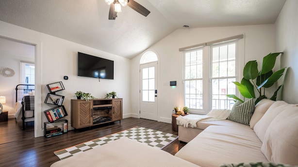 A spacious living room with a large sofa, a television mounted on the wall, and a wooden cabinet with books and decorative items. - Core's Cottage: Fountain Square- Cozy 1BR Dwtn Indy (Indianapolis)