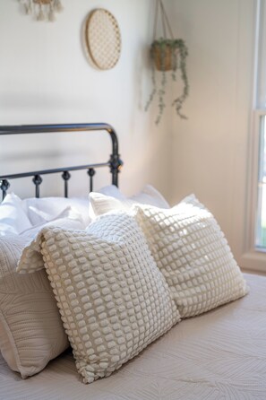Una cama con ropa blanca y almohadas texturizadas, un tapiz tejido y una planta colgante.