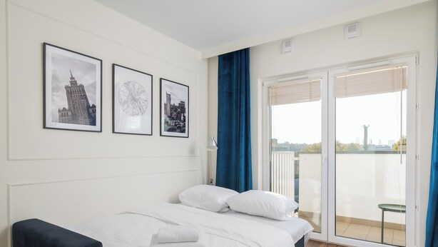 A bedroom with a bed, a nightstand, a window with a view of buildings, and three framed pictures on the wall. - Praga Bliska Glamour Studio (Warszawa)