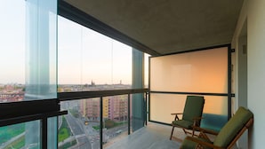 Un pis a un edifici alt amb grans finestres del sostre al terra, dues cadires verdes amb coixins i vista a un paisatge urbà amb edificis i una carretera.