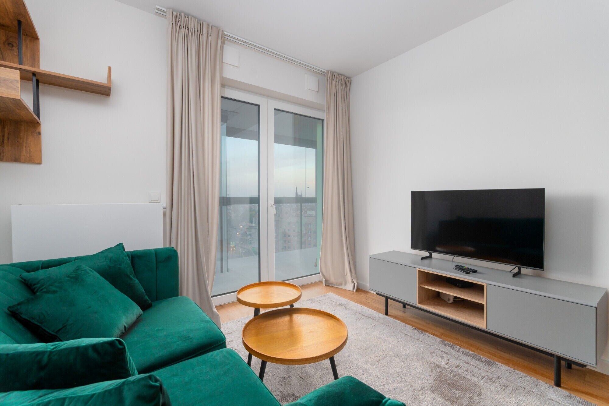 Ein modernes Wohnzimmer mit einem grünen Sofa, zwei Holznachttischen, einer grauen Unterhaltungsecke mit Flachbildfernseher und einem großen Fenster mit Vorhängen.
