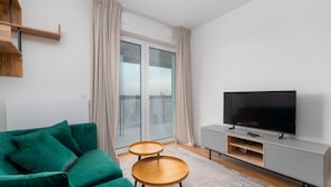 Un saló modern amb un sofà verd, dues taules laterals de fusta, un moble de televisió gris amb un televisor de pantalla plana, i una finestra gran amb cortines.