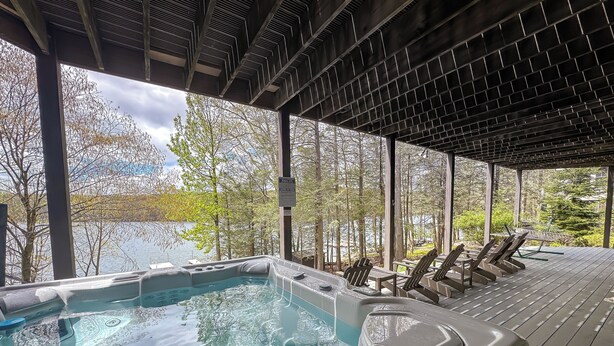 Et spabad med udsigt til en sø og træer, omgivet af træterrasse stole og en pergola.