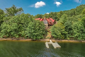 A lakeside cabin with a red roof, surrounded by dense forest, and accessible by a wooden dock. - The Iconic Red Roof Lakefront Estate of Deep Creek Lake - 8BR, Sleeps 20 (McHenry)