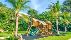 A wooden playground structure with slides and climbing elements, surrounded by palm trees and greenery. - Stunning 4BR Luxury Retreat w Private Rooftop Pool (Playa Del Carmen)