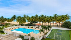 Zona de piscina con cancha de baloncesto, rodeada de palmeras y una playa.