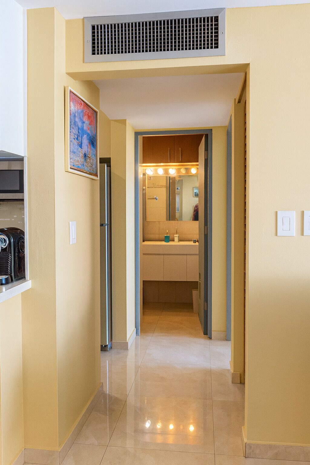 A hallway leading to a kitchen with a refrigerator, a sink, and a mirror with lights.