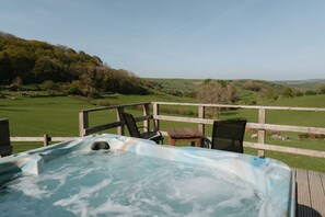 A hot tub on a wooden deck with a view of rolling hills and a wooden fence.