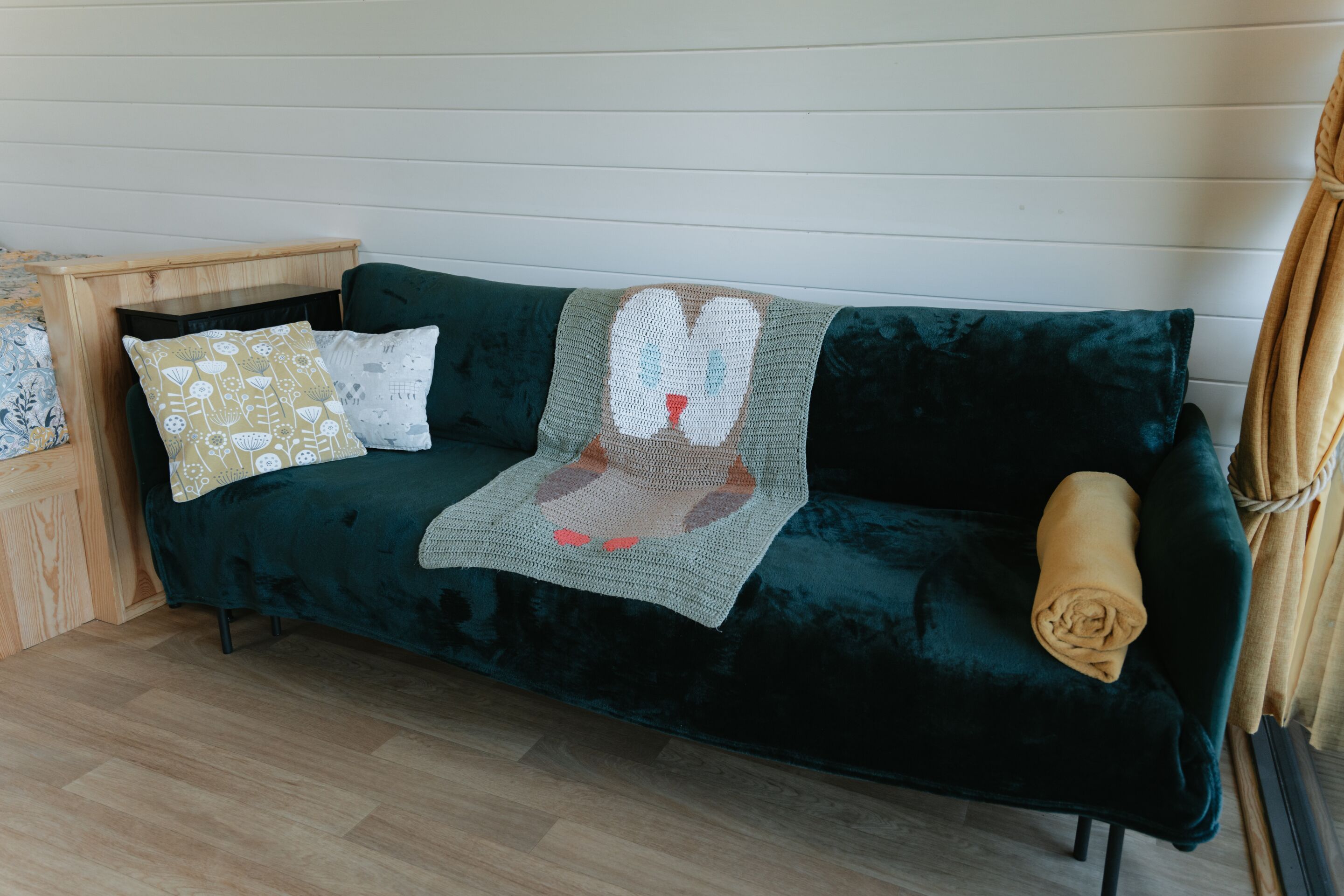 A dark green sofa with a blanket and two pillows, a wooden side table, and a rolled-up blanket on the floor.