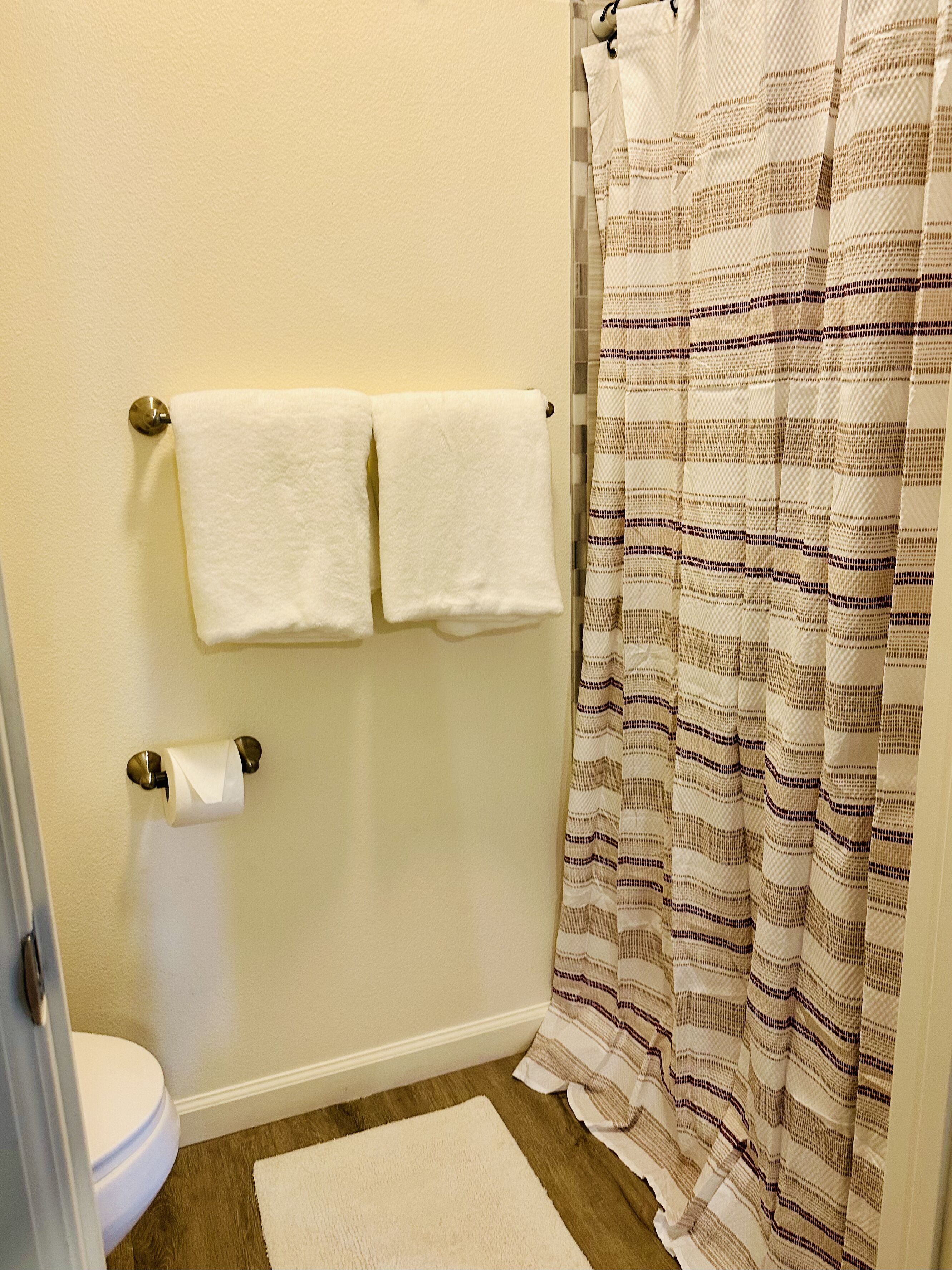 Ein Badezimmer mit Toilette, zwei Handtüchern an einem Ablagegestell, einer Duschvorhang und einem kleinen Teppich auf dem Boden.