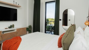 Een moderne hotelkamer met een groot bed, een flatscreen televisie, een spiegel en een balkon met uitzicht op groen.